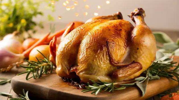 Frango assado com pele dourada e crocante, rodeado por ervas frescas e vegetais assados.