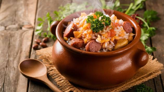 Preparando arroz, feijão e linguiça na panela de barro com ingredientes frescos ao fundo.